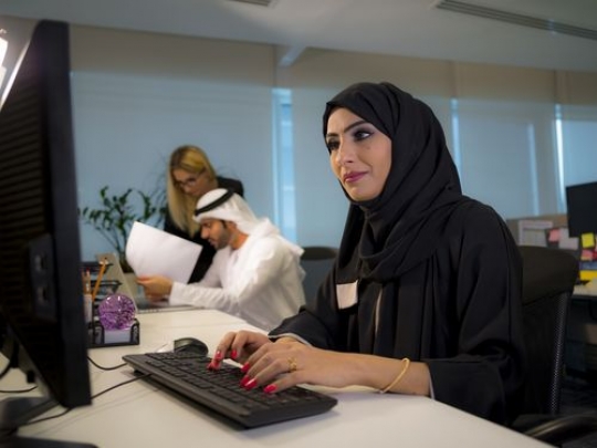 Arab business woman sitting by her desk with a background of two people working together|
