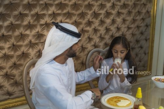 Young Arab girl drinking milk sitting next to her father during breakfast|