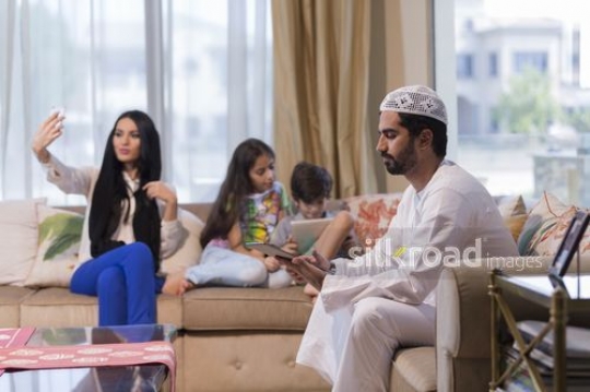 Middle Eastern woman taking a photo of herself with the young kids and man are on their device|