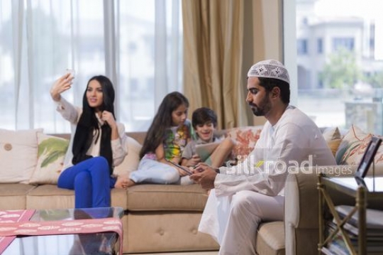 Middle Eastern man and young kids sitting with their devices while the woman takes a selfie |