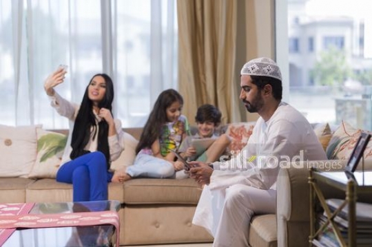 Middle Eastern man and young kids on their devices while the Middle Eastern woman is taking a selfie|