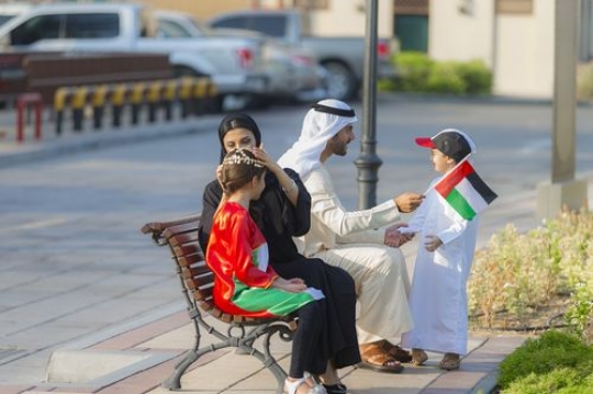 Mother helping her daughter wear the traditional head jewellery while the father is carrying a UAE flag and talking to his son|-