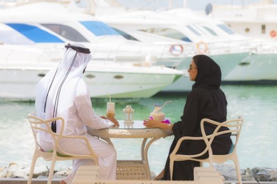 Man and woman dressed in the traditional UAE attire while sitting outside talking to each other|