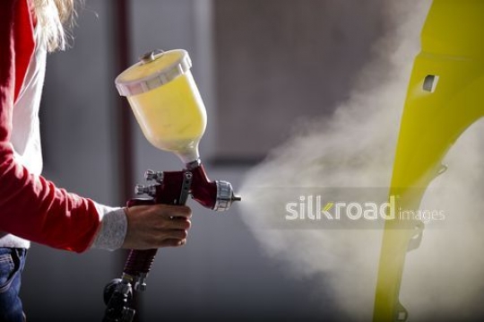 Woman spraying paint on car part|-